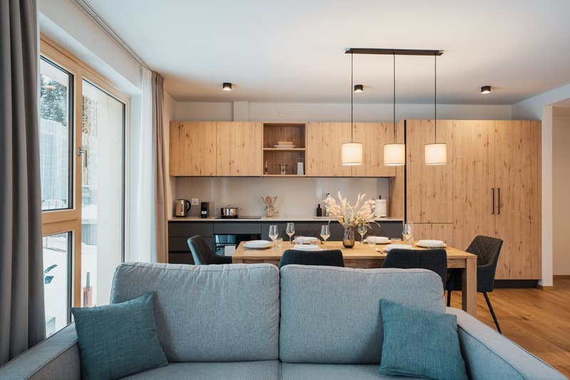 The image shows an open-concept living room and kitchen area. The kitchen features wooden cabinets and shelves, a black countertop, and pendant lighting above the dining table. The living room has a large gray sofa and accent pillows. There is a sliding g
