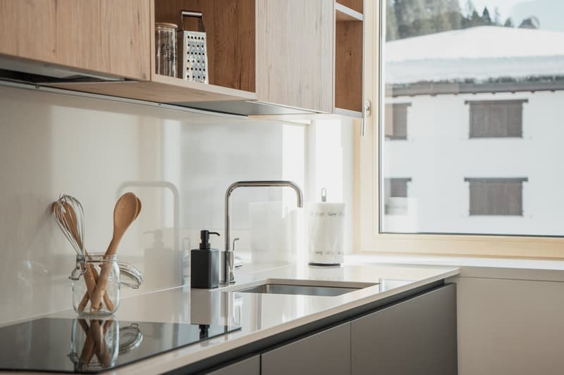 Modern kitchen with wooden cabinets, stainless steel appliances, and a large window overlooking the outdoors. The kitchen features a sink with a faucet, a dishwasher, and various cooking utensils and accessories stored in the cabinets.