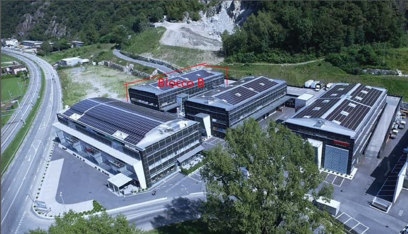 Large multi-story commercial building with solar panels on the roof, surrounded by parking lots and greenery. The building appears to be a modern industrial or office complex located in a mountainous area.