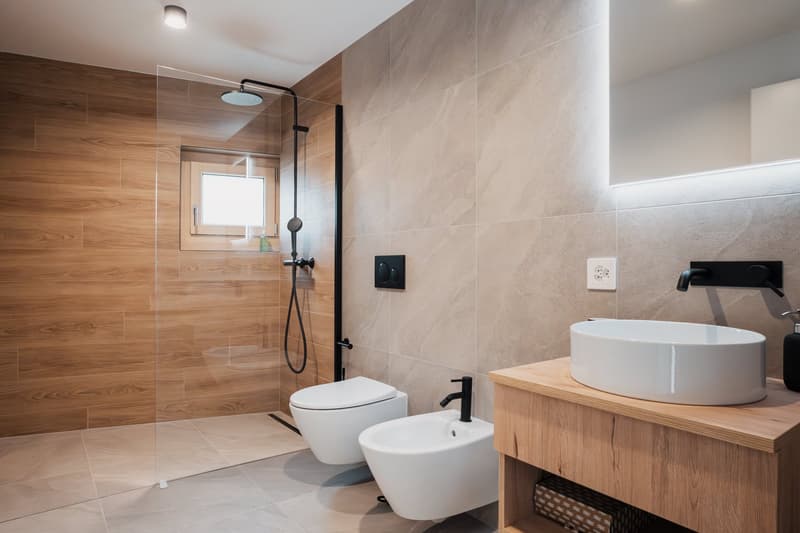 Modern bathroom with wood-paneled walls, large walk-in shower, white toilet and bidet, wooden vanity with vessel sink, and a window