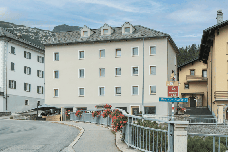 multistory beige building, large windows, flowers, iron fences, gray roof, street sign, stone pavement, stairs, mountain background