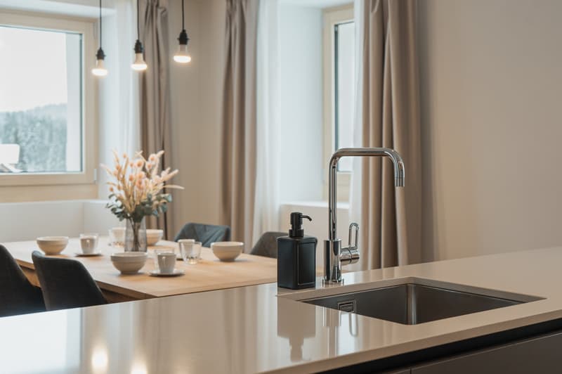 Modern kitchen with dining area, granite countertops, stainless steel sink, wooden dining table, dining chairs, kitchen faucet with a bottle of hand soap, a dining table with place settings, and a window with a view outside