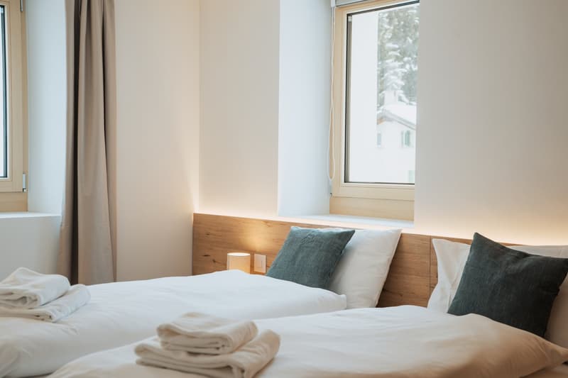 Two beds, pillows, bedside lamp, windows with curtains