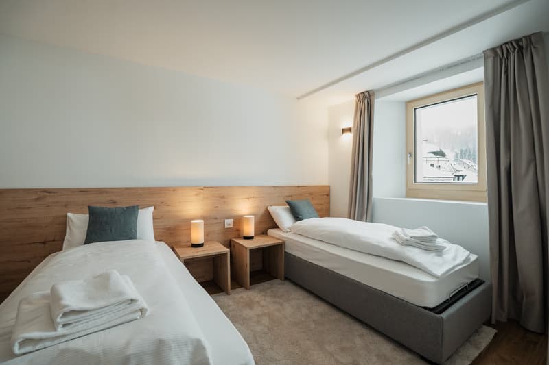 Bedroom with two twin beds, lamps, side tables, gray headboard, white walls, carpet, large window with view of snowy mountains