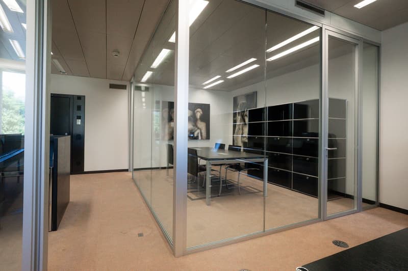 Modern office space with glass walls, a conference table, and artwork on the walls. The room has a dishwasher visible, indicating it may have a kitchenette or break area.