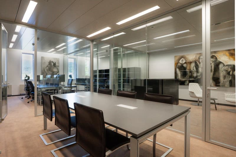 Large open office space with glass walls, modern furniture, and artwork on the walls. The room has a long conference table with leather chairs, and there are additional workstations visible through the glass walls.