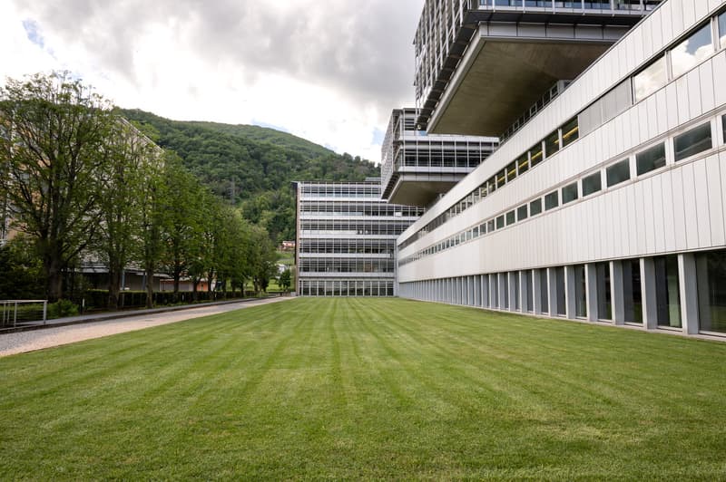 A large modern office building with multiple floors, surrounded by a well-maintained lawn and trees. The building has a sleek, glass and concrete design with a raised ground floor and visible parking spaces on the side.