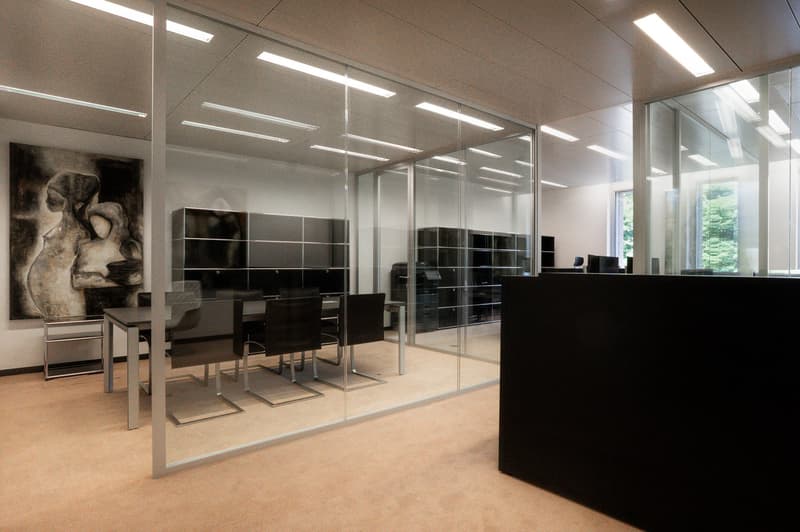 Large open office space with glass walls, black furniture and storage units, and a large abstract artwork on the wall.