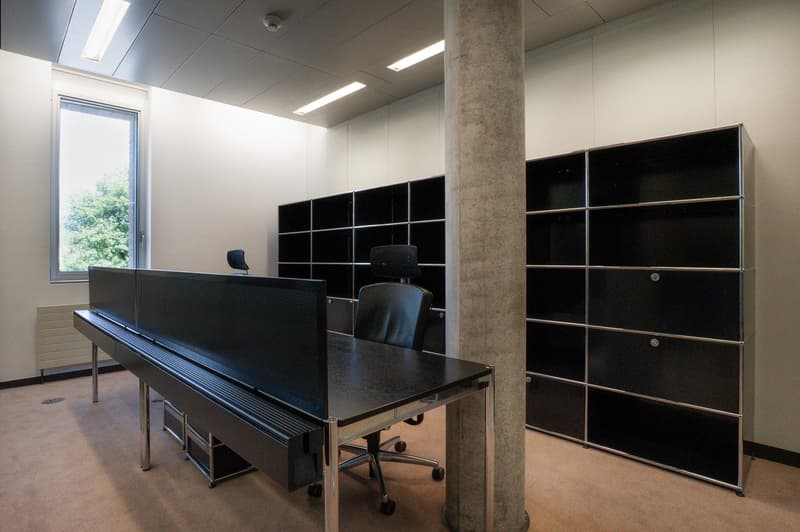 The image shows an office space with a large black desk, black office chairs, and black storage cabinets along the walls. The room has a concrete column and large windows providing natural light. The overall design is minimalist and modern.