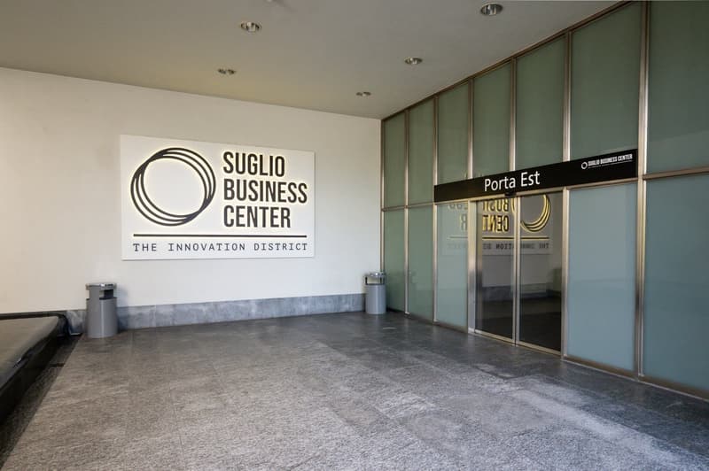 The image shows the lobby of the Suglio Business Center, which is described as 'The Innovation District'. The lobby has a large logo sign on the wall, glass doors leading to the exterior, and a concrete floor. The space appears to be a modern and minimali