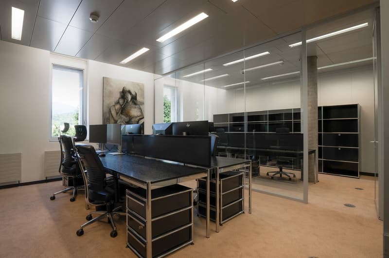 The image shows a modern and well-lit office space. The room has a large central table with several black office chairs around it, and there are multiple computer workstations along the walls. The space is illuminated by overhead lighting fixtures and has