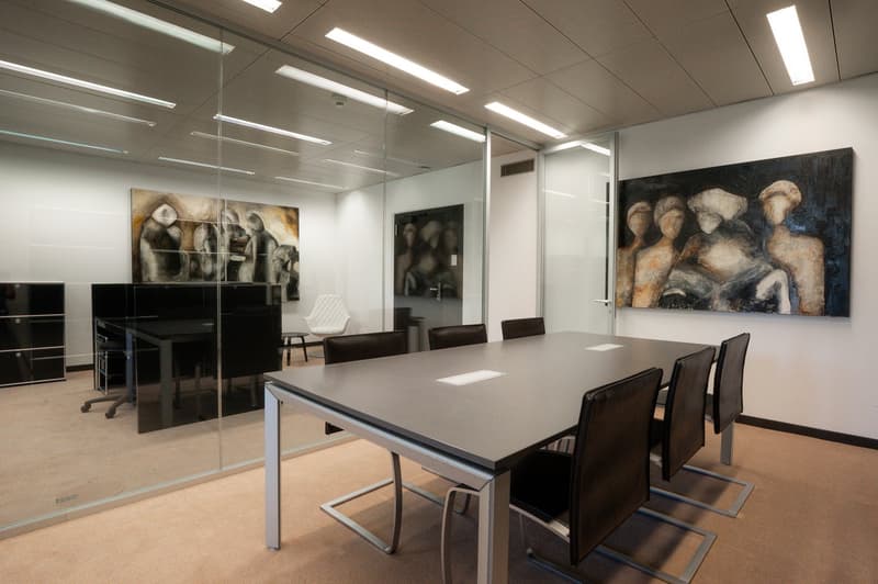 Large modern meeting room with a long gray table, black leather chairs, and abstract artwork on the walls. The room has ample lighting from the ceiling fixtures and a glass wall separating it from the rest of the office space.