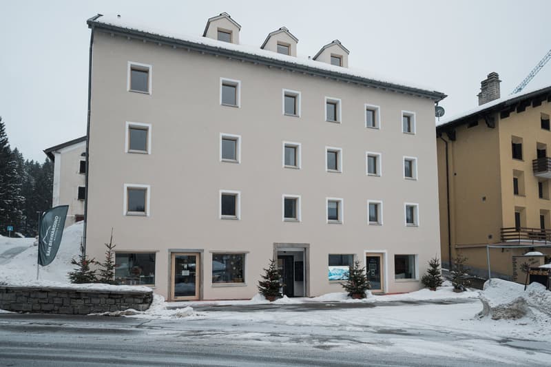 Exterior view of a four-story building with a flat roof, covered in snow. The building has numerous windows, a small balcony on the upper floor, and decorative plants outside the main entrance. Adjacent buildings and a snow-covered hill are visible in the