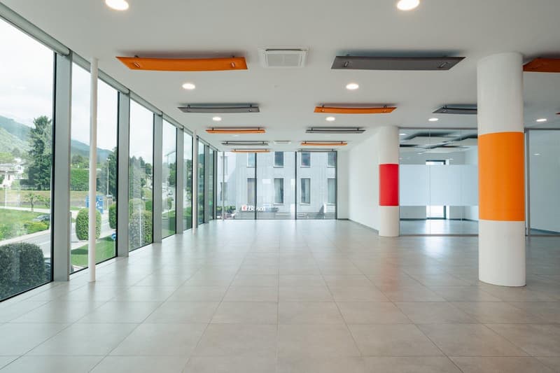 A spacious building hallway with large glass windows, tiled floor, tall columns, and bright ceiling lighting.
