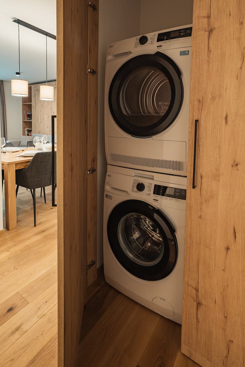 A room with a stack of washing machine and dishwasher, wooden floor, and a dining table with chairs.