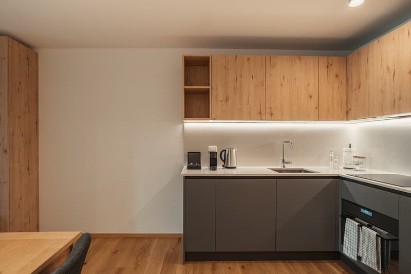Modern kitchen with wooden cabinets, white countertops, and a wooden floor. Equipped with a sink, faucet, electric kettle, coffee machine, and various kitchen items.