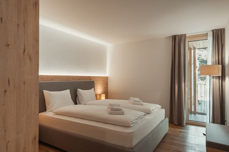 A bedroom with a bed, pillows, and blankets. There are towels on the bed, a lamp on the nightstand, and a curtain on the window.