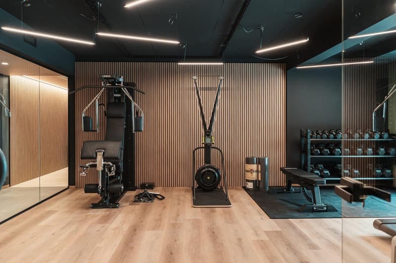 Modern gym with weight machines, dumbbells, and a bench. Wooden flooring and vertical wooden wall panels.
