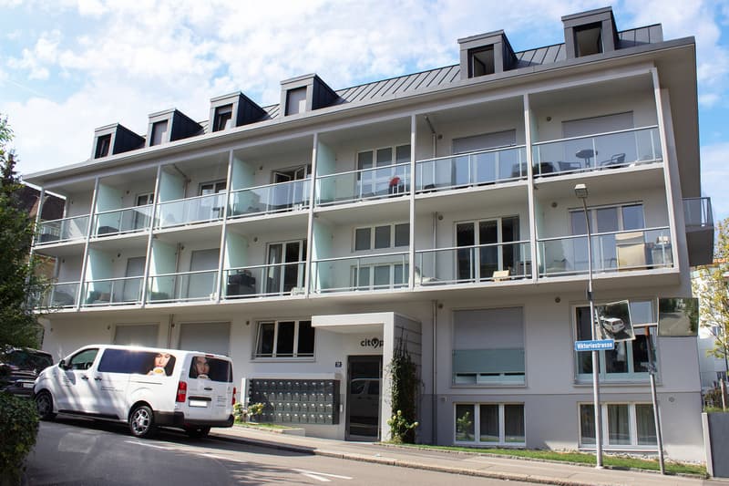 Multi-story apartment building with modern, minimalist design. The building has several balconies and a parking area in front. The exterior features a combination of glass, concrete, and metal elements.
