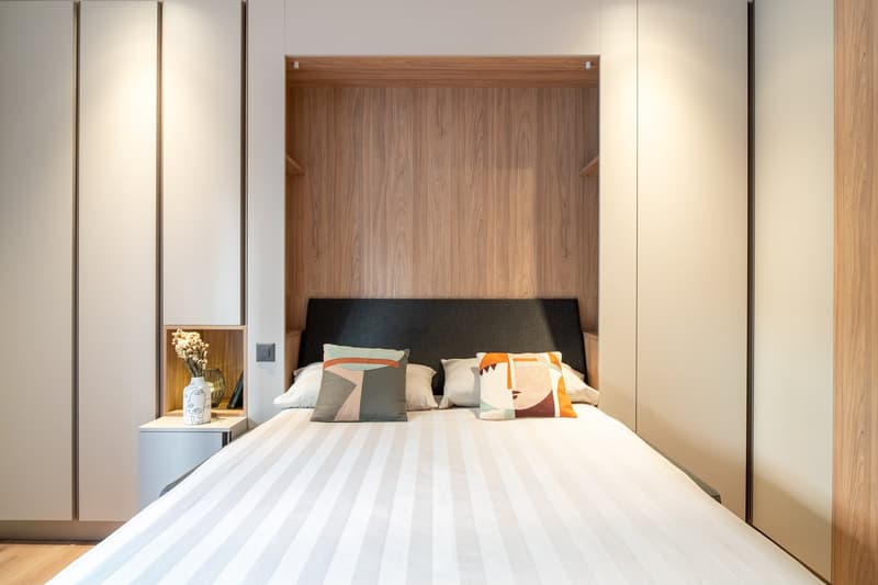 Spacious bedroom with a wooden headboard, white bedding, and decorative pillows. The room features built-in wardrobes on the side walls and a nightstand with a vase of flowers.