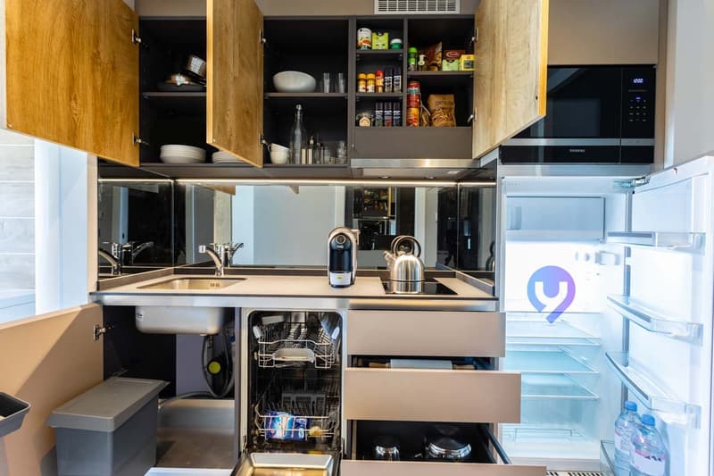 Modern kitchen with wooden cabinets, stainless steel appliances including a dishwasher and microwave, and a refrigerator. The kitchen has a sink and countertop space, as well as storage shelves filled with various kitchen items and supplies.