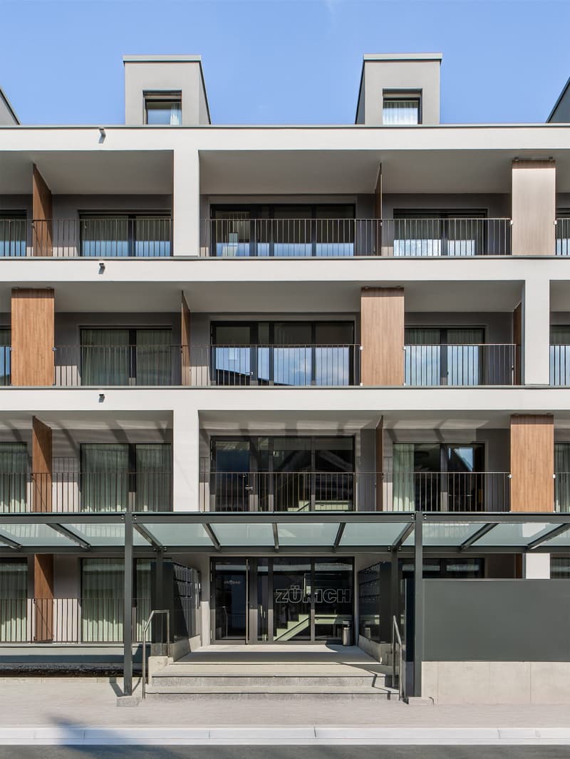 Multi-story building with a modern, minimalist design. The building has a glass and concrete facade with balconies on each floor. The entrance is marked by the 'Zurich' sign.