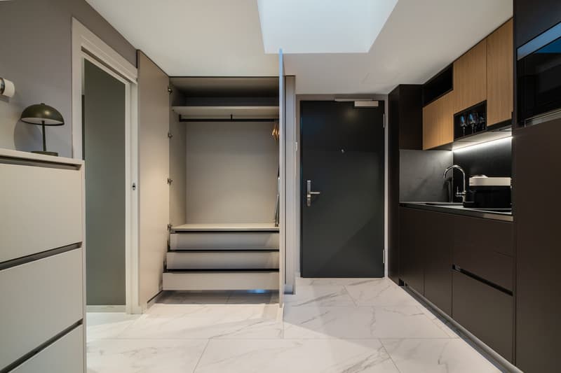 Open-concept studio apartment with a kitchenette, built-in closet, and marble-like flooring. The apartment features a dark color scheme with black cabinets and accents, complemented by lighter walls and floors. A dishwasher is visible in the kitchenette a