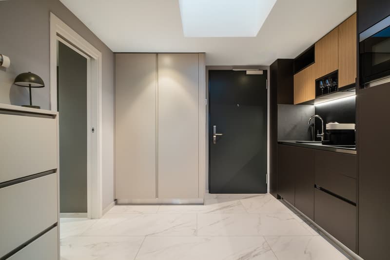 Modern and minimalist apartment interior with open-plan kitchen and living area. The space features a marble-like tile flooring, built-in kitchen cabinets with a dark finish, and a dishwasher. The entrance door and closet doors are also visible.