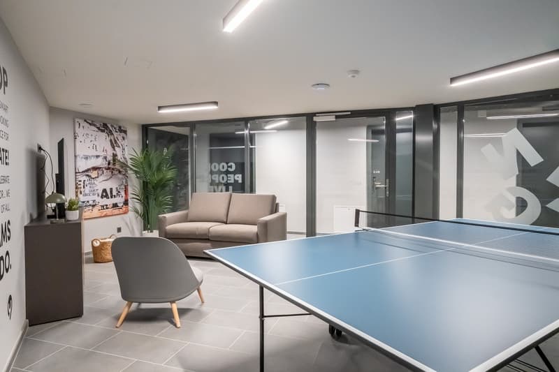 The image shows a modern and stylish office space with a ping pong table in the center. The room has a minimalist design with white walls, gray tile flooring, and large windows. There is a comfortable sofa and a potted plant, creating a relaxed and inviti