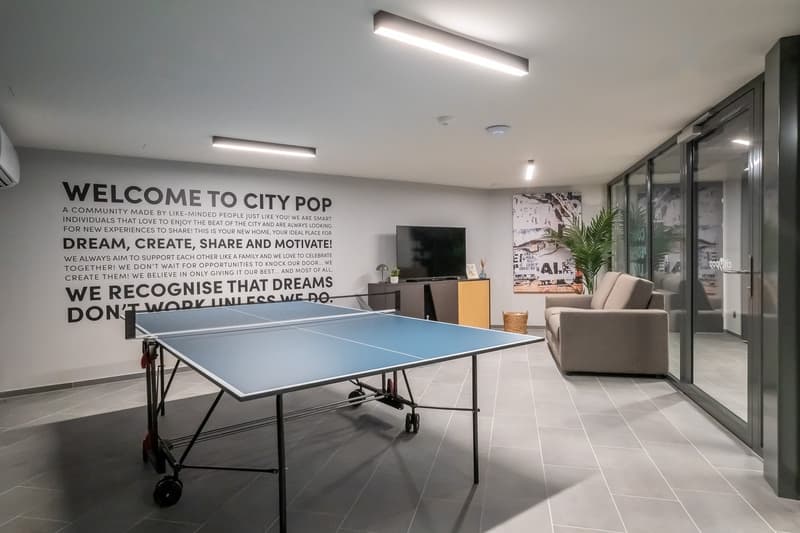 The image shows a modern office space with a ping pong table in the center. The walls feature a large motivational message that encourages dreaming, creating, sharing, and motivating. The space also includes comfortable seating, storage units, and a tropi