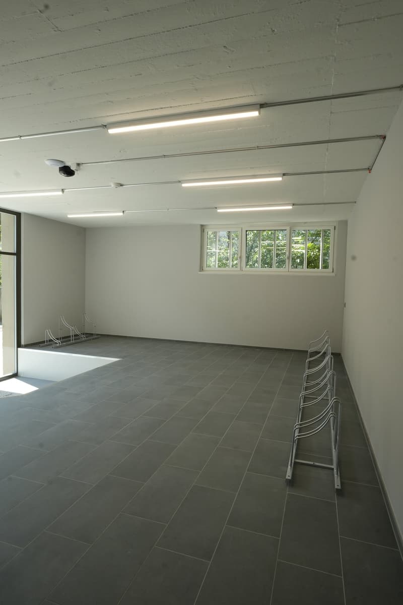 Large open room with gray tile flooring, white walls, and multiple fluorescent light fixtures on the ceiling. The room has several windows overlooking a wooded outdoor area. There are several chairs lined up along the walls, suggesting this may be a meeti