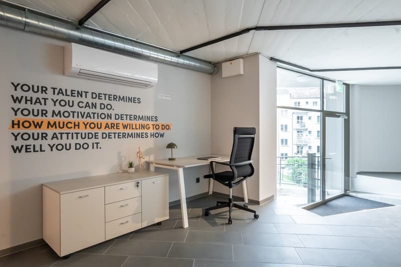 Modern office space with large windows, white walls, gray tile flooring, a desk with a computer, and a motivational quote displayed on the wall.