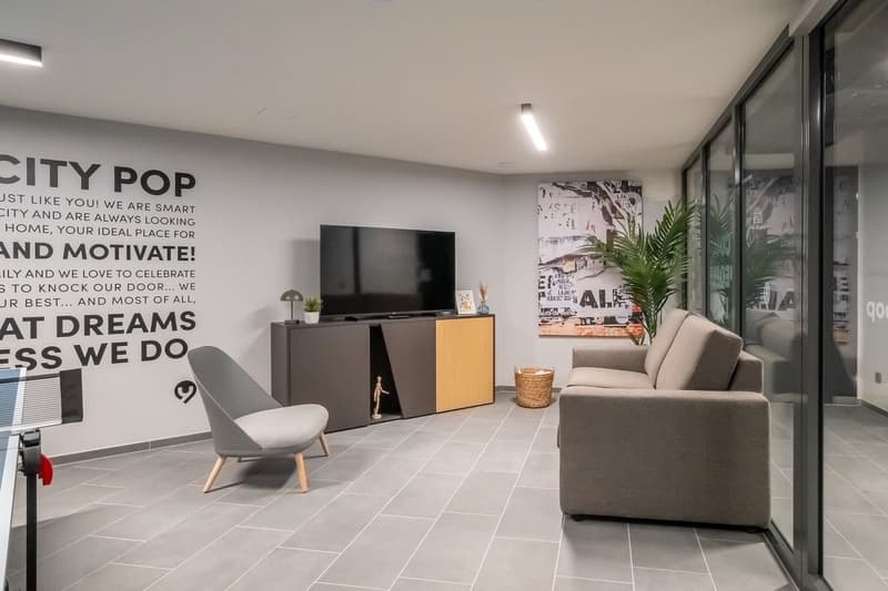 Modern living room with large windows, gray tile flooring, gray sofa, TV stand with TV, and a motivational wall mural