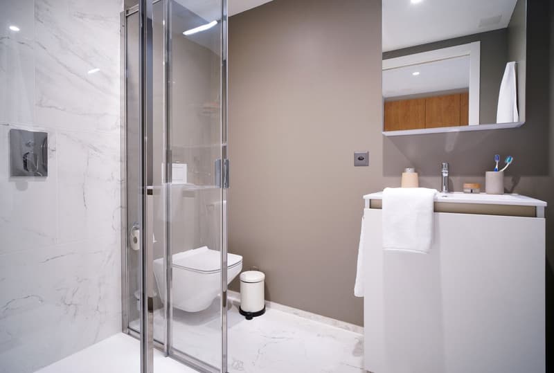 Modern bathroom with marble-like tiles, glass shower enclosure, vanity with sink, and a toilet. The bathroom also has a private washing machine visible.