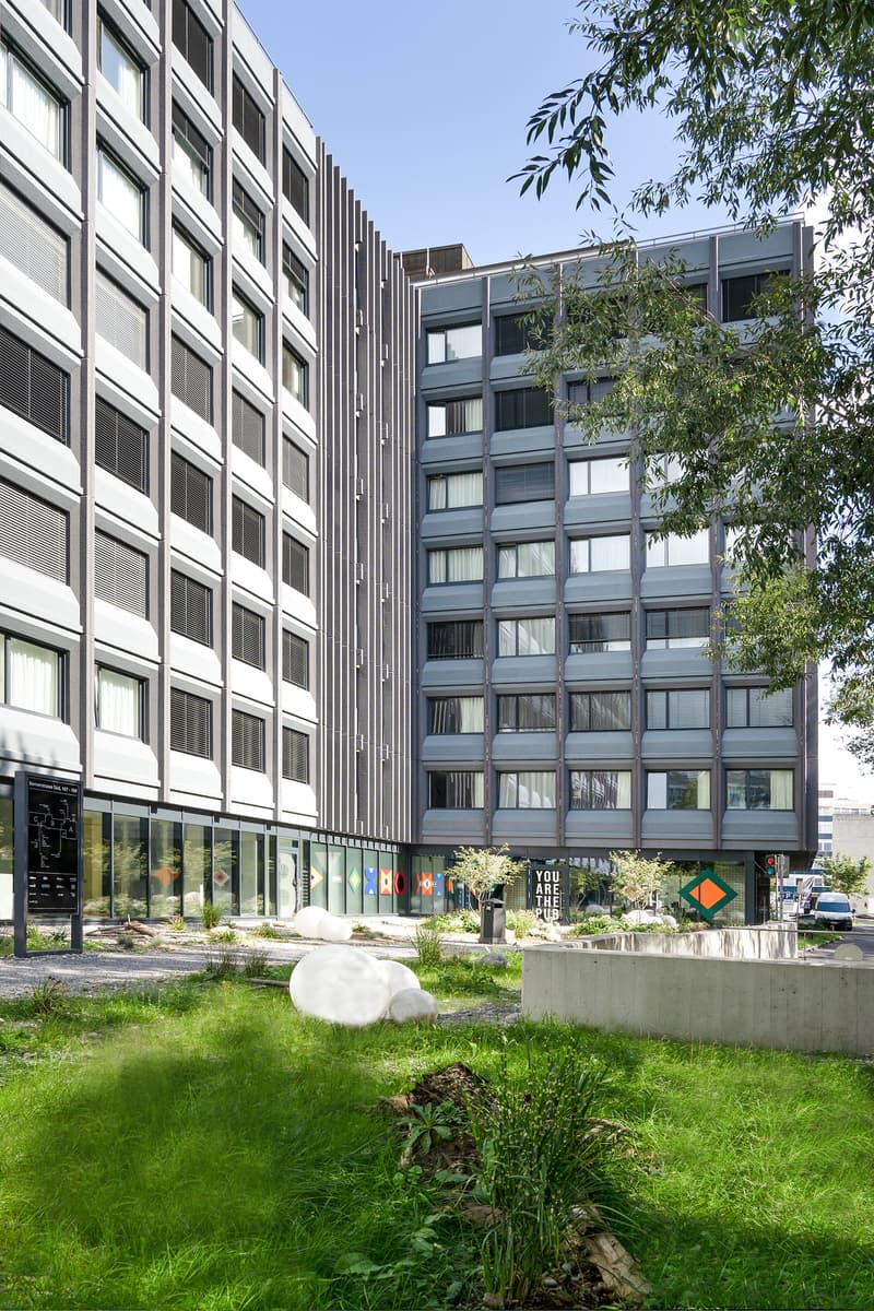 Multi-story office building with a modern, geometric facade, surrounded by greenery and landscaping. The building has large windows, a ground floor with retail or commercial spaces, and a grassy area with sculptures or art installations in the foreground.