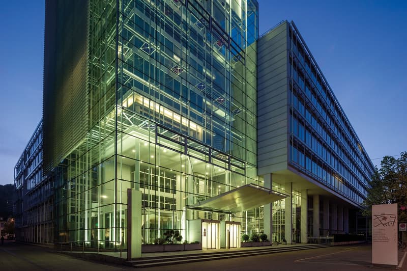 Large modern glass and steel office building with a prominent entrance and parking area in front. The building has multiple floors and a sleek, contemporary design with a lot of natural light coming through the glass facade.