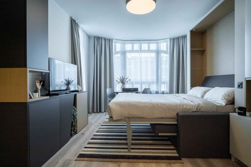 Spacious bedroom with large bay windows, gray curtains, wooden flooring, a king-size bed, a TV stand, and a striped area rug.