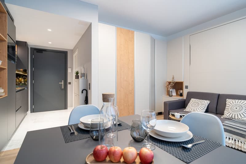 The image shows a modern and stylish open-plan living and dining area. The space features a gray sofa, a dining table with place settings, and a kitchen area with a dishwasher visible. The walls are painted in light colors, and the room has a mix of woode