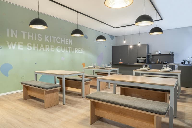 Modern and open kitchen space with wooden tables and benches, black pendant lights, and a large wall text that says 'In this kitchen we share cultures'.