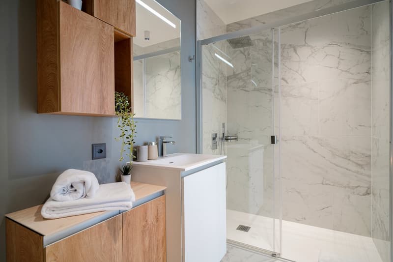 Modern bathroom with marble-like tile walls, glass shower enclosure, vanity with white sink and wooden cabinets, towels, and a potted plant
