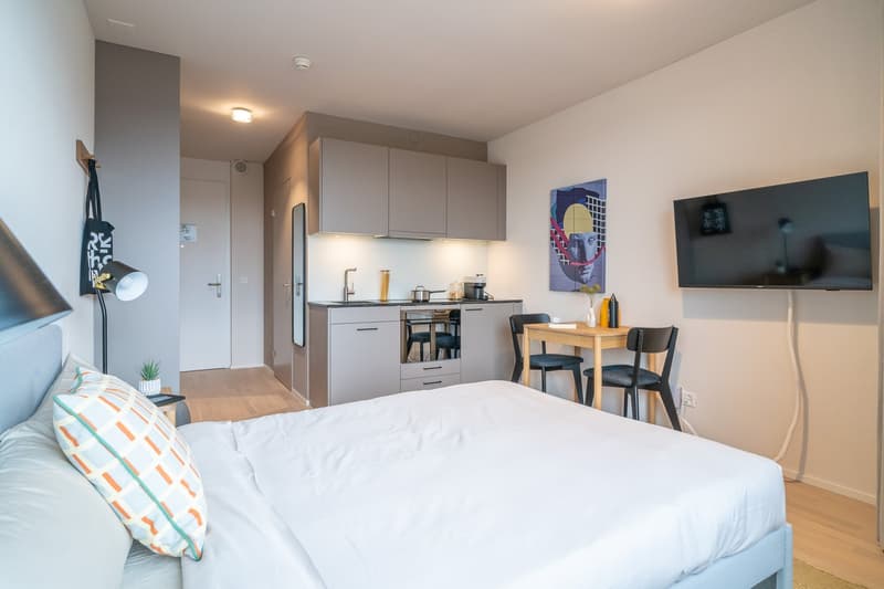 The image shows a studio apartment with an open-plan layout. The room features a bed, a small dining area with a table and chairs, and a kitchenette with cabinets, a stove, and a dishwasher. The walls are painted in neutral tones, and there is artwork and