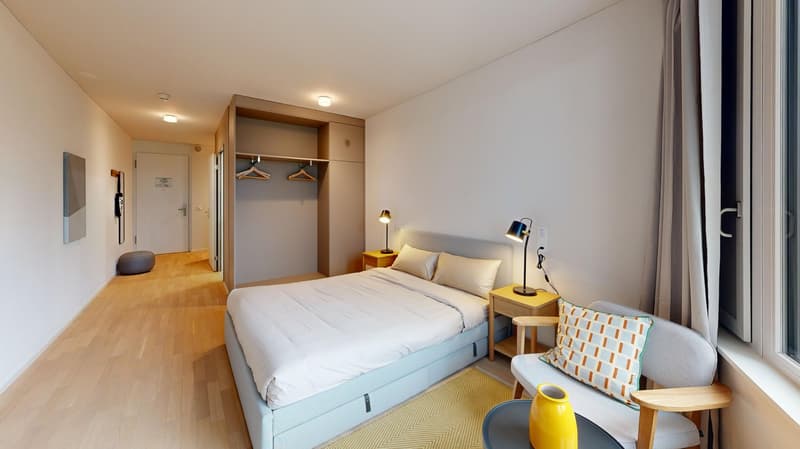 Spacious bedroom with a large bed, wooden wardrobe, bedside tables, and a comfortable chair. The room has large windows and a balcony or terrace area.