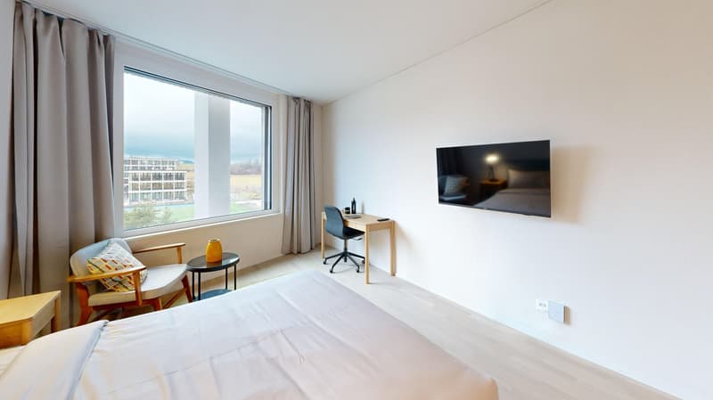 Spacious bedroom with large window providing a view, white walls, hardwood floor, bed, desk, and TV mounted on the wall