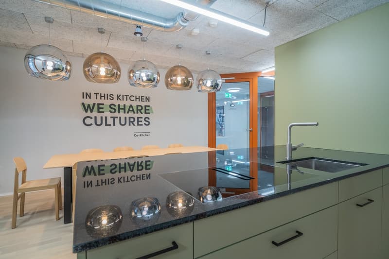 Modern kitchen with pendant lighting, large table, and text on the wall stating 'In this kitchen we share cultures'