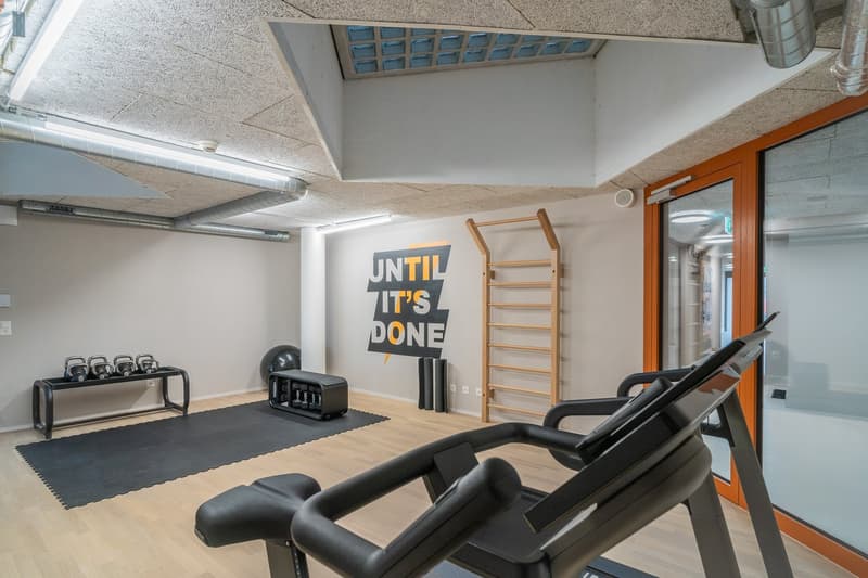 The image shows a gym or fitness room with various exercise equipment, including a ladder on the wall. The room has a motivational wall decal that says 'Until it's done'. The room has a hardwood floor and exposed ceiling with ventilation ducts.