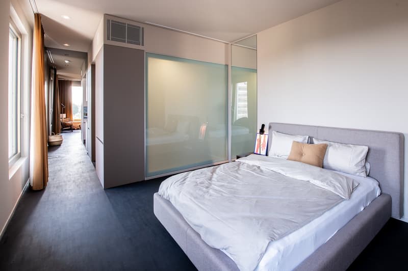 Spacious bedroom with a large bed, gray upholstered headboard, and a sliding glass door leading to a balcony or terrace. The room has a minimalist and modern design with white walls and dark hardwood floors.