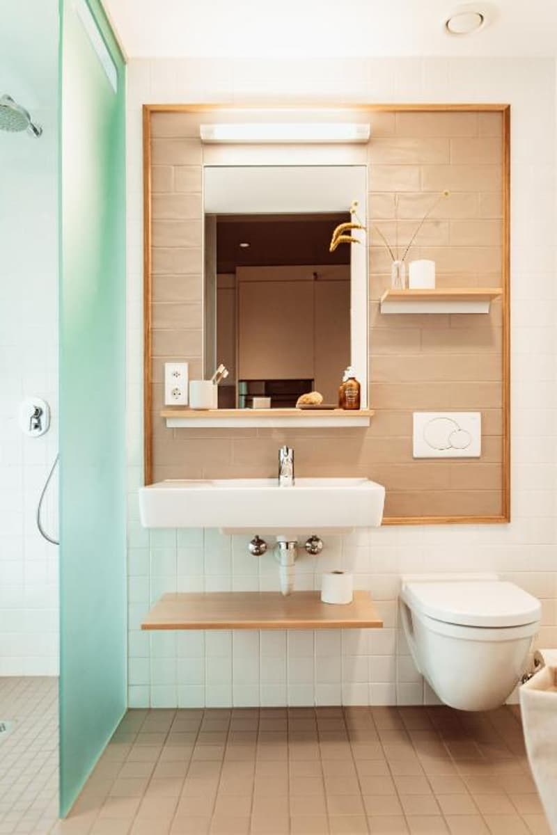 The image shows a modern bathroom with a white sink, a mirror with a wooden frame, and shelves for storage. The bathroom has a clean and minimalist design with neutral colors and natural materials like wood.