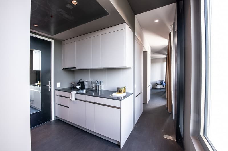Modern kitchen with white cabinets, black countertops, and stainless steel appliances including a dishwasher. The kitchen leads to a hallway with dark flooring.