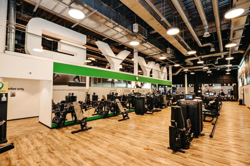 Large open gym space with various workout equipment such as weight machines and cardio machines, wooden flooring, high ceilings with exposed ductwork, and bright lighting.