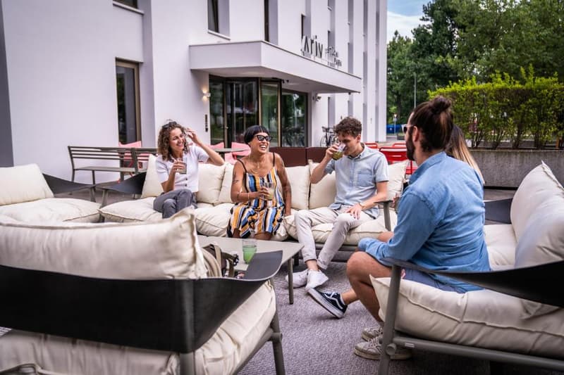 The image shows a group of young people sitting on comfortable outdoor furniture, such as sofas and chairs, in an outdoor seating area. They appear to be socializing and enjoying drinks. The setting is in front of a modern building with large windows.